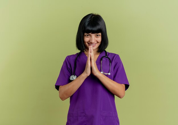 Glimlachende jonge brunette vrouwelijke arts in uniform met stethoscoop houdt handen bij elkaar geïsoleerd op olijfgroene achtergrond met kopie ruimte