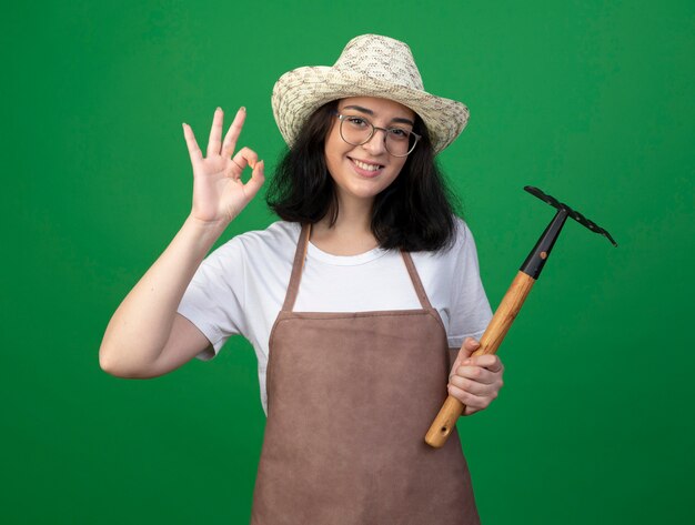 Glimlachende jonge brunette vrouwelijke tuinman in optische bril en uniform dragen tuinieren hoed gebaren ok handteken en houdt hark geïsoleerd op groene muur