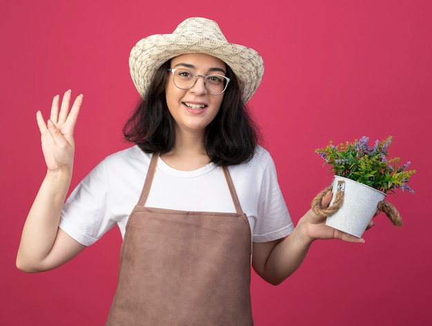 Glimlachende jonge brunette vrouwelijke tuinman in optische bril en uniform dragen tuinieren hoed houdt bloempot en gebaren vier met vingers geïsoleerd op roze muur met kopie ruimte