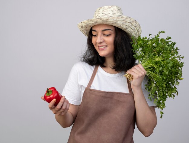 Glimlachende jonge brunette vrouwelijke tuinman in uniform dragen tuinieren hoed houdt koriander en kijkt naar rode peper geïsoleerd op een witte muur