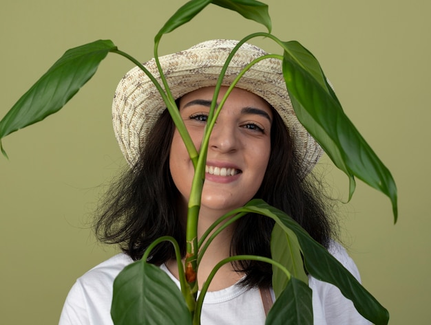 Gratis foto glimlachende jonge brunette vrouwelijke tuinman in uniform dragen tuinieren hoed staat achter plant geïsoleerd op olijfgroene muur