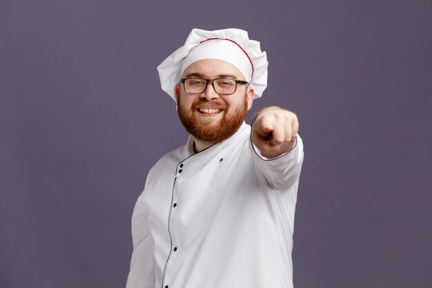 Glimlachende jonge chef-kok met een uniforme bril en pet kijken en wijzend op camera geïsoleerd op paarse achtergrond