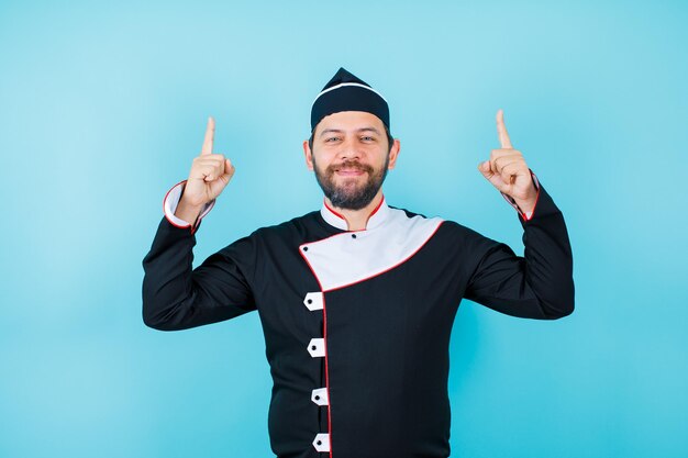 Glimlachende jonge chef-kok wijst omhoog met wijsvingers op blauwe achtergrond
