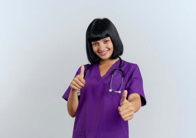 Glimlachende jonge donkerbruine vrouwelijke arts in uniform met stethoscoop beduimelt omhoog met twee handen