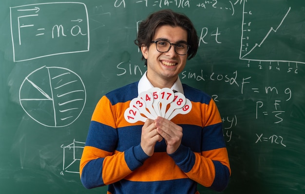 glimlachende jonge geometrieleraar met een bril die voor het schoolbord staat in de klas met nummerfans die naar voren kijken