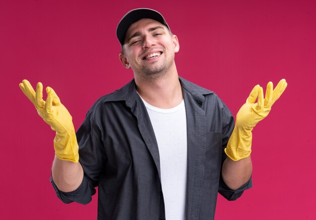 Glimlachende jonge hamdsome schoonmaakster die t-shirt en pet met handschoenen spreidt die handen spreiden die op roze muur worden geïsoleerd