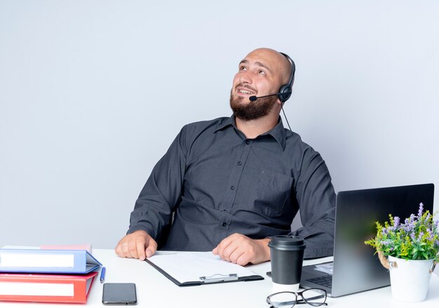 Glimlachende jonge kale callcentermens die hoofdtelefoonszitting bij bureau met uitrustingsstukken dragen die omhoog geïsoleerd op witte muur kijken