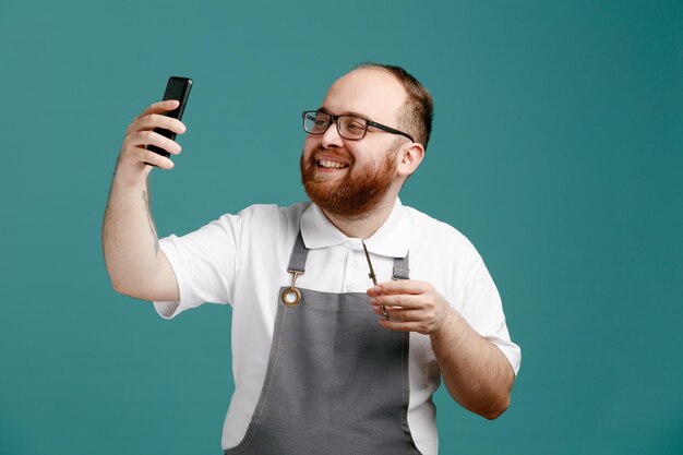 Glimlachende jonge kapper in uniform en bril met schaar die selfie neemt met mobiele telefoon geïsoleerd op blauwe achtergrond