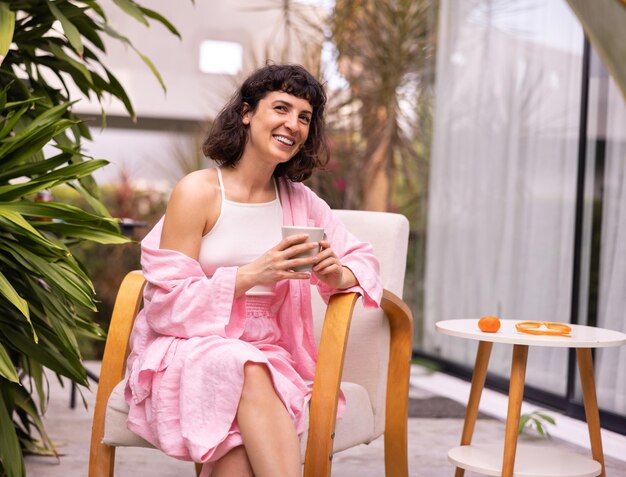 Glimlachende jonge kaukasische brunette vrouw zit op een stoel met een kopje koffie in de ochtend in de achtertuin. Ontspanningsconcept