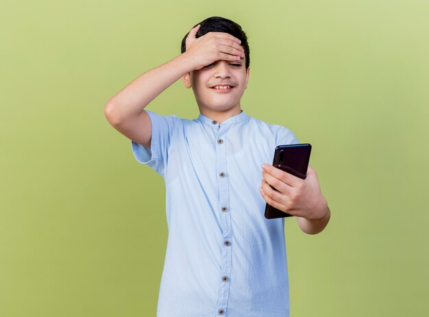 Glimlachende jonge Kaukasische jongen die en mobiele telefoon houdt bekijkt die hand op voorhoofd houdt dat op olijfgroene achtergrond met exemplaarruimte wordt geïsoleerd