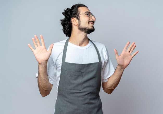 Glimlachende jonge Kaukasische mannelijke kapper die glazen en golvende haarband draagt ?? in uniform die lege handen toont die kant bekijken die op witte achtergrond wordt geïsoleerd