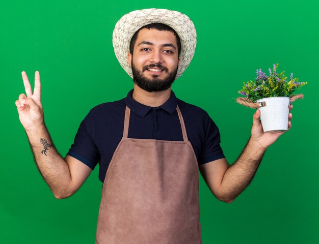 Glimlachende jonge kaukasische mannelijke tuinman die een tuinierhoed draagt die bloempot houdt en een overwinningsteken gebaart dat op groene muur met exemplaarruimte wordt geïsoleerd