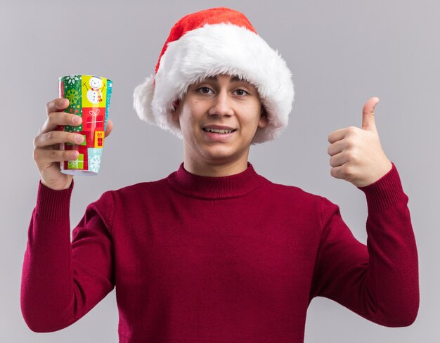 Glimlachende jonge kerel die Kerstmishoed draagt die Kerstmiskop houdt die duim toont die omhoog op witte muur wordt geïsoleerd