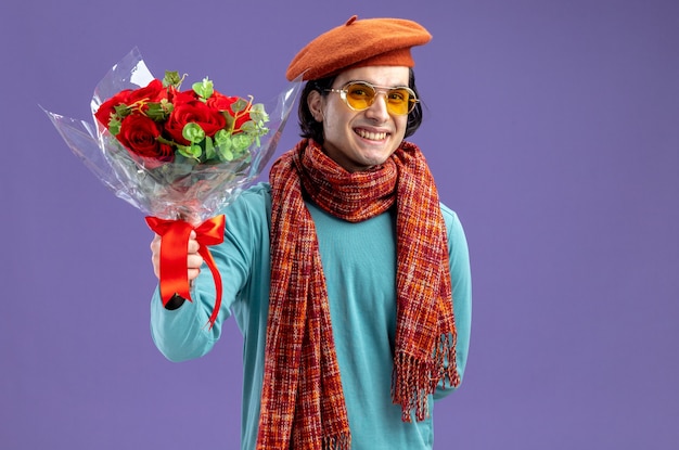 Gratis foto glimlachende jonge kerel op valentijnsdag met hoed met sjaal en bril die boeket standhoudt op camera geïsoleerd op blauwe achtergrond