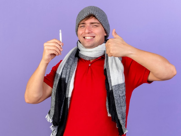 Glimlachende jonge knappe blonde zieke man die de winterhoed en sjaal draagt die thermometer houdt die voorzijde toont die duim toont die omhoog op purpere muur wordt geïsoleerd