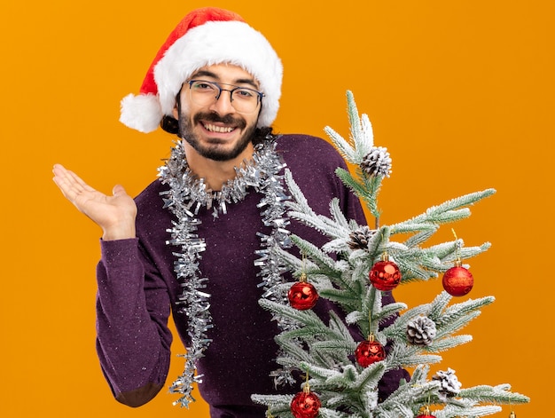 Glimlachende jonge knappe kerel die achter de kerstboom staat en een kerstmuts draagt met een slinger op de nek die de hand spreidt geïsoleerd op een oranje muur