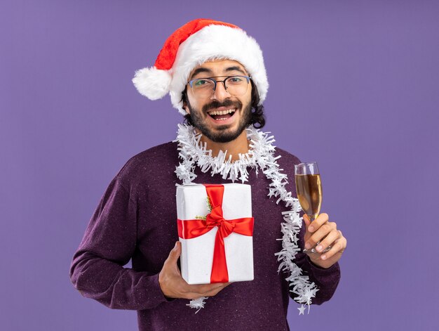 Glimlachende jonge knappe kerel die Kerstmishoed met slinger op hals draagt die giftdoos met glas champagne houdt die op blauwe achtergrond wordt geïsoleerd