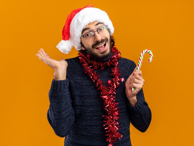 Glimlachende jonge knappe kerel die Kerstmishoed met slinger op hals draagt die Kerstmissuikergoed verspreiden die hand op oranje achtergrond wordt geïsoleerd