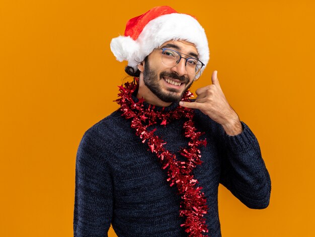 Glimlachende jonge knappe kerel die Kerstmishoed met slinger op hals draagt die telefoongesprekgebaar toont dat op oranje achtergrond wordt geïsoleerd