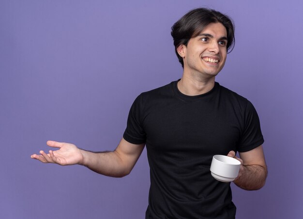 Glimlachende jonge knappe kerel die zwarte t-shirt draagt die kop van koffie houdt die hand spreidt die op purpere muur wordt geïsoleerd