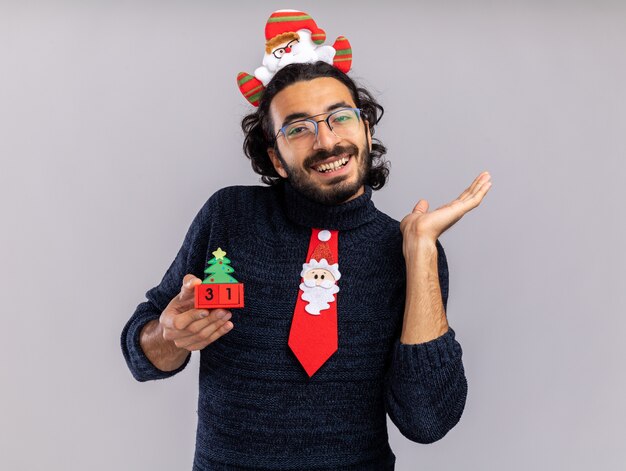 Glimlachende jonge knappe kerel met kerststropdas met haarhoepel die kerstspeelgoedpunten vasthoudt met de hand aan de zijkant geïsoleerd op een witte muur met kopieerruimte