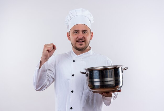 Glimlachende jonge knappe kok in de pot van de chef-kok eenvormige holding en het opheffen van vuist die op witte ruimte wordt geïsoleerd