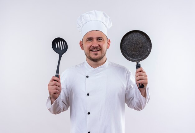 Glimlachende jonge knappe kok in eenvormige chef-kok met ingelaste lepel en koekenpan op geïsoleerde witte ruimte