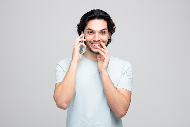 Glimlachende jonge knappe man die de hand in de buurt van de mond houdt en naar de camera kijkt terwijl hij aan de telefoon praat op een witte achtergrond