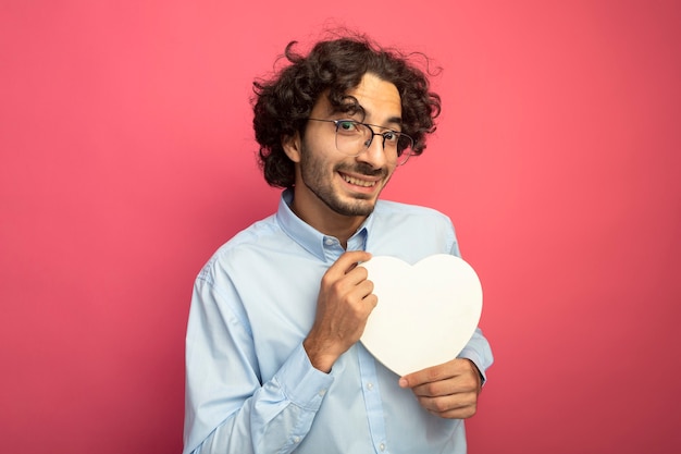 Glimlachende jonge knappe man die een bril draagt die hartvorm houdt die voorzijde bekijkt die op roze muur wordt geïsoleerd