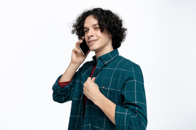 Glimlachende jonge knappe man die in profielweergave staat en naar de zijkant kijkt terwijl hij aan de telefoon praat terwijl hij zijn shirt grijpt dat op een witte achtergrond wordt geïsoleerd