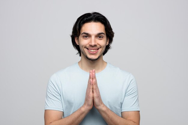 Glimlachende jonge knappe man die naar camera kijkt die namaste-gebaar toont dat op witte achtergrond wordt geïsoleerd