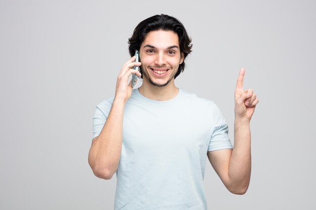 Glimlachende jonge knappe man die naar de camera kijkt en omhoog wijst terwijl hij aan de telefoon praat op een witte achtergrond