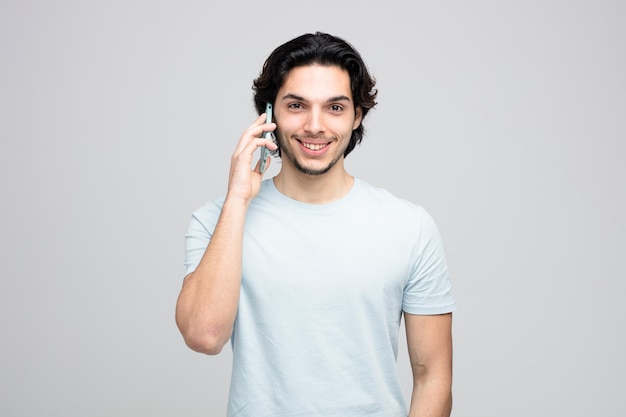 Glimlachende jonge knappe man die naar de camera kijkt terwijl hij aan de telefoon praat op een witte achtergrond