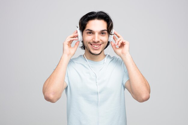 Glimlachende jonge knappe man met koptelefoon die ze aanraakt en kijkt naar camera geïsoleerd op een witte achtergrond