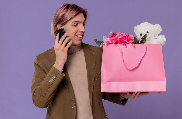 Glimlachende jonge knappe man praten aan de telefoon en kijken naar roze cadeauzakje met bloemen en teddybeer