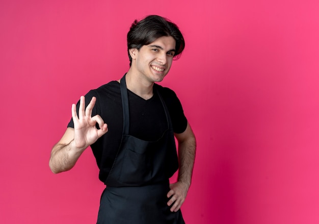 Glimlachende jonge knappe mannelijke kapper in uniform hand op heup zetten en okey gebaar geïsoleerd op roze met kopie ruimte tonen