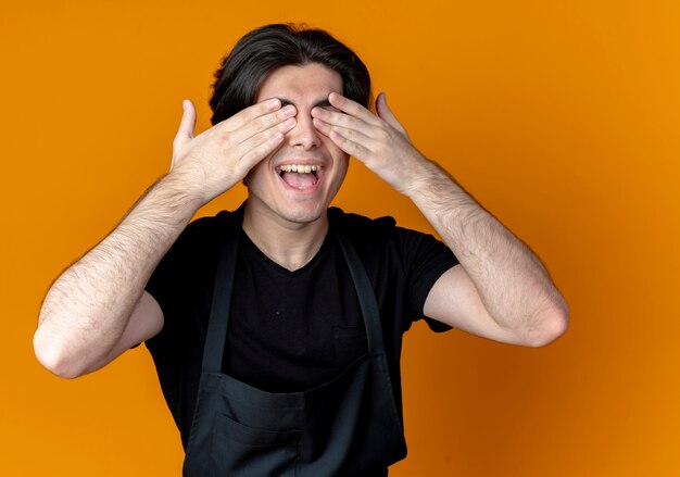 Glimlachende jonge knappe mannelijke kapper in uniforme sluitende ogen met handen geïsoleerd op oranje