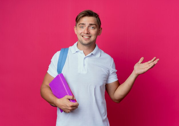 Glimlachende jonge knappe mannelijke student die de boeken en de punten van de achterzakholding met hand aan kant draagt die op roze muur wordt geïsoleerd