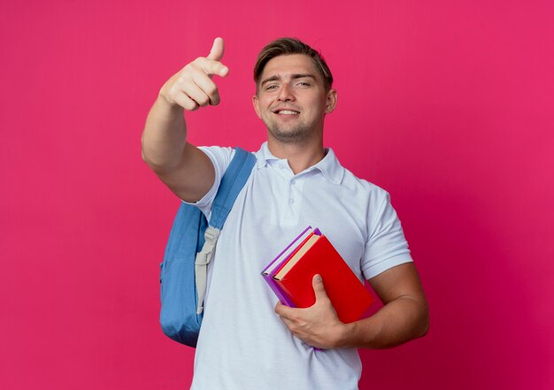 Glimlachende jonge knappe mannelijke student die de boeken en punten van de achterzakholding draagt die op roze muur worden geïsoleerd