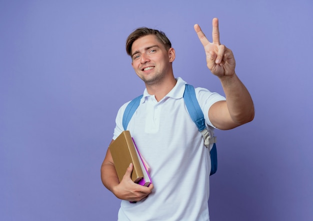 Glimlachende jonge knappe mannelijke student die de boeken van de achterzakholding draagt en vredesgebaar toont dat op blauw wordt geïsoleerd