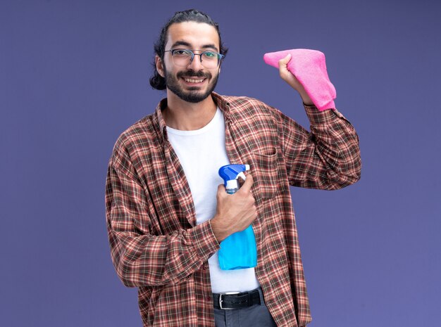 Glimlachende jonge knappe schoonmaakster die een t-shirt draagt met een spuitfles met een vod geïsoleerd op een blauwe muur