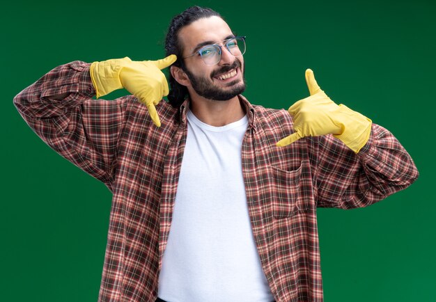 Glimlachende jonge knappe schoonmaakster met een t-shirt en handschoenen met een telefoongebaar geïsoleerd op een groene muur