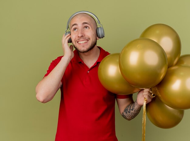 Glimlachende jonge knappe Slavische partijkerel die hoofdtelefoons draagt die ballons houden die hoofdtelefoons aanraken die omhoog geïsoleerd op olijfgroene achtergrond kijken