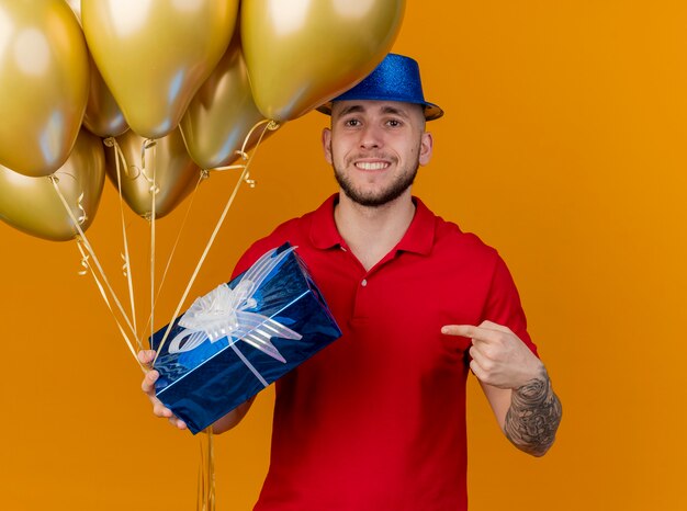 Glimlachende jonge knappe Slavische partijkerel die partijhoed draagt die ballons en giftpak houdt die camera bekijkt die op giftpak richt dat op oranje achtergrond wordt geïsoleerd