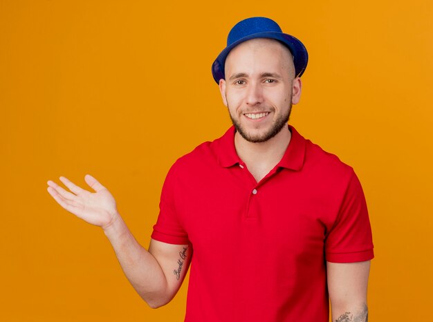 Glimlachende jonge knappe Slavische partijkerel die partijhoed draagt die camera bekijkt die lege die hand op oranje achtergrond toont