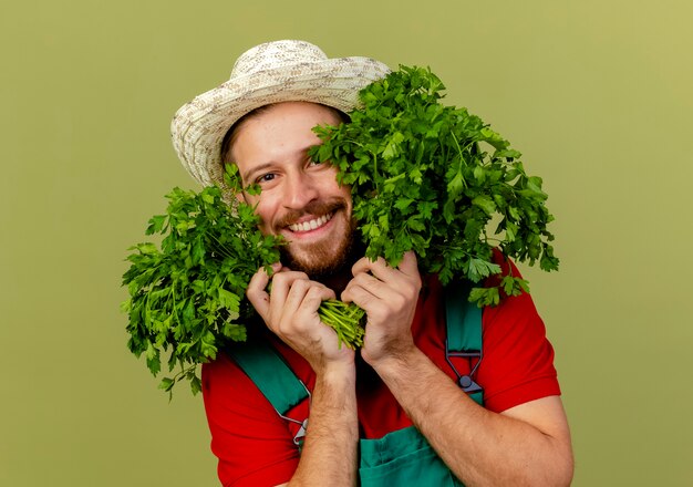 Glimlachende jonge knappe Slavische tuinman in uniform en hoed met korianders in de buurt van gezicht geïsoleerd op olijfgroene muur