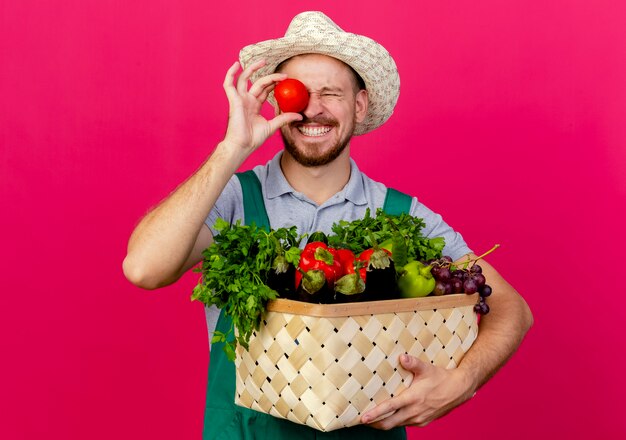 Glimlachende jonge knappe slavische tuinman in uniform en hoed met mand met groenten met tomaat op oog met gesloten ogen geïsoleerd op karmozijnrode muur