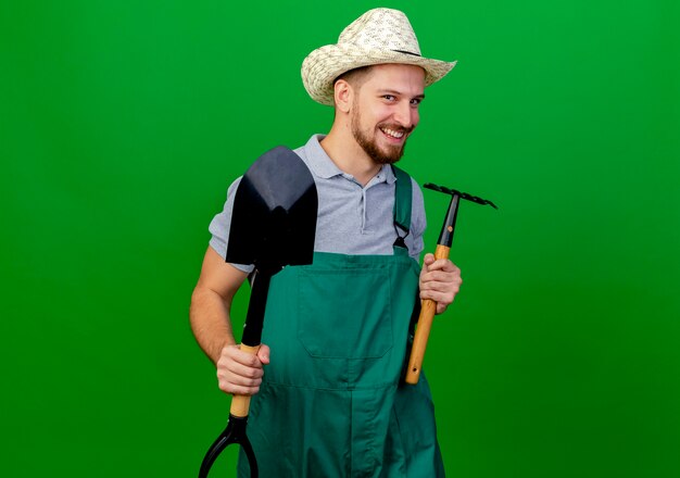 Glimlachende jonge knappe Slavische tuinman in uniform en hoed op zoek met hark en schop