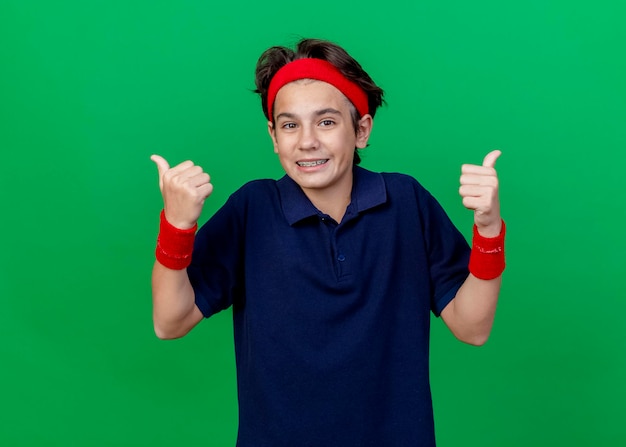 Glimlachende jonge knappe sportieve jongen die hoofdband en polsbandjes met beugels draagt die aan de voorkant kijken duimen opdagen geïsoleerd op groene muur met kopie ruimte