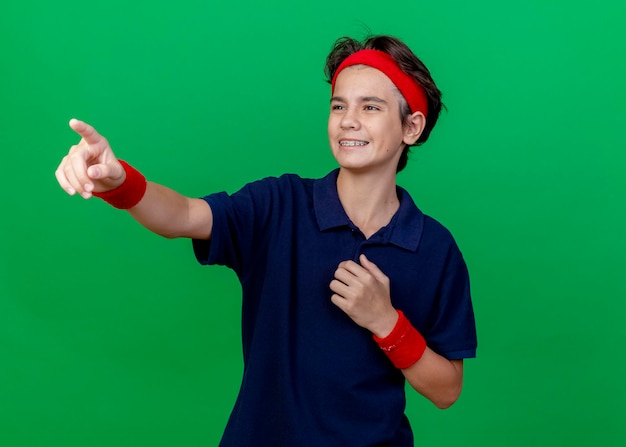 Glimlachende jonge knappe sportieve jongen die hoofdband en polsbandjes met beugels draagt ?? die borst aanraken kijken en wijzen naar kant geïsoleerd op groene muur met kopie ruimte
